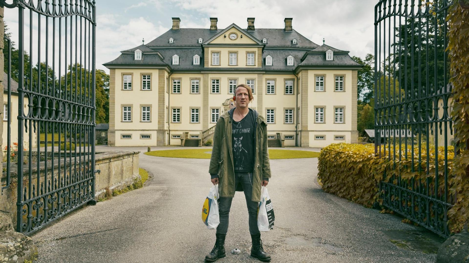 Szenenfoto aus "Happy Burnout". Wotan Wilke Möhring als "Fussel" steht in Punkkleidung und mit Plastiktüten vor dem exklusiven Sanatorium (Foto: Thomas Kost / Riva Film)