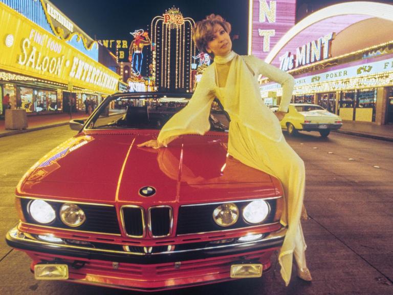 Moderatorin Carolin Reiber 1982 in Las Vegas bei den Dreharbeiten für eine Silvestershow. Sie posiert auf einem roten BMW-Sportwagen, im Hintergrund sind die Lichter der Casinos zu sehen.