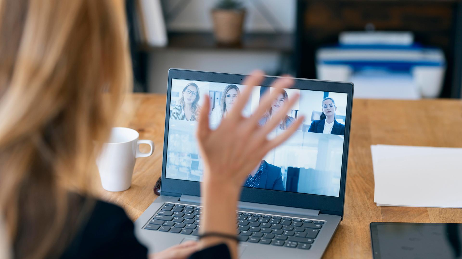 Eine Frau hebt beim Videocall in Home Office ihre Hand und winkt den Teilnehmern am Bildschirm zu. Eine Frau hebt beim Videocall in Home Office ihre Hand und winkt den Teilnehmern am Bildschirm zu.