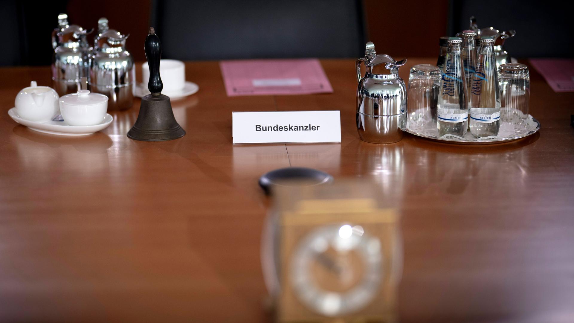 Ein Holztisch im Kanzleramt ist gedeckt mit Tassen, Gläsern, Wasserfläschchen und kleinen Kannen. Auf dem Tisch steht ein Aufsteller mit "Bundeskanzler".