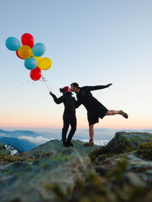 Eine Frau mit Luftballons in der Hand und ein Mann, der ihr einen Kuss gibt. Beide stehen auf der Spitze eines Berges. Eine Frau mit Luftballons in der Hand und ein Mann, der ihr einen Kuss gibt. Beide stehen auf der Spitze eines Berges.