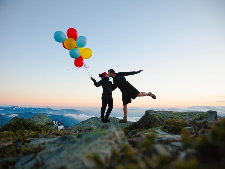 Eine Frau mit Luftballons in der Hand und ein Mann, der ihr einen Kuss gibt. Beide stehen auf der Spitze eines Berges. Eine Frau mit Luftballons in der Hand und ein Mann, der ihr einen Kuss gibt. Beide stehen auf der Spitze eines Berges.