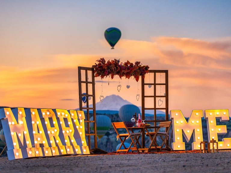 Große leuchtende Schriftzeichen "Marry me" stehen am Strand, inklusive Holrahmen und dekoriertem Tisch mit Stühlen