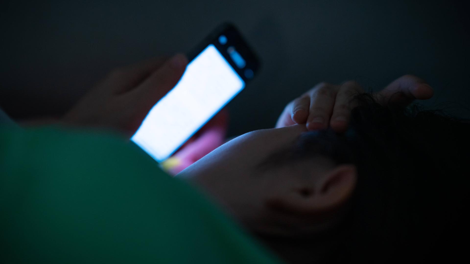 Foto einer jungen Frau, die im Dunkeln im Bett liegt und auf ihr blaues Smartphone-Display guckt