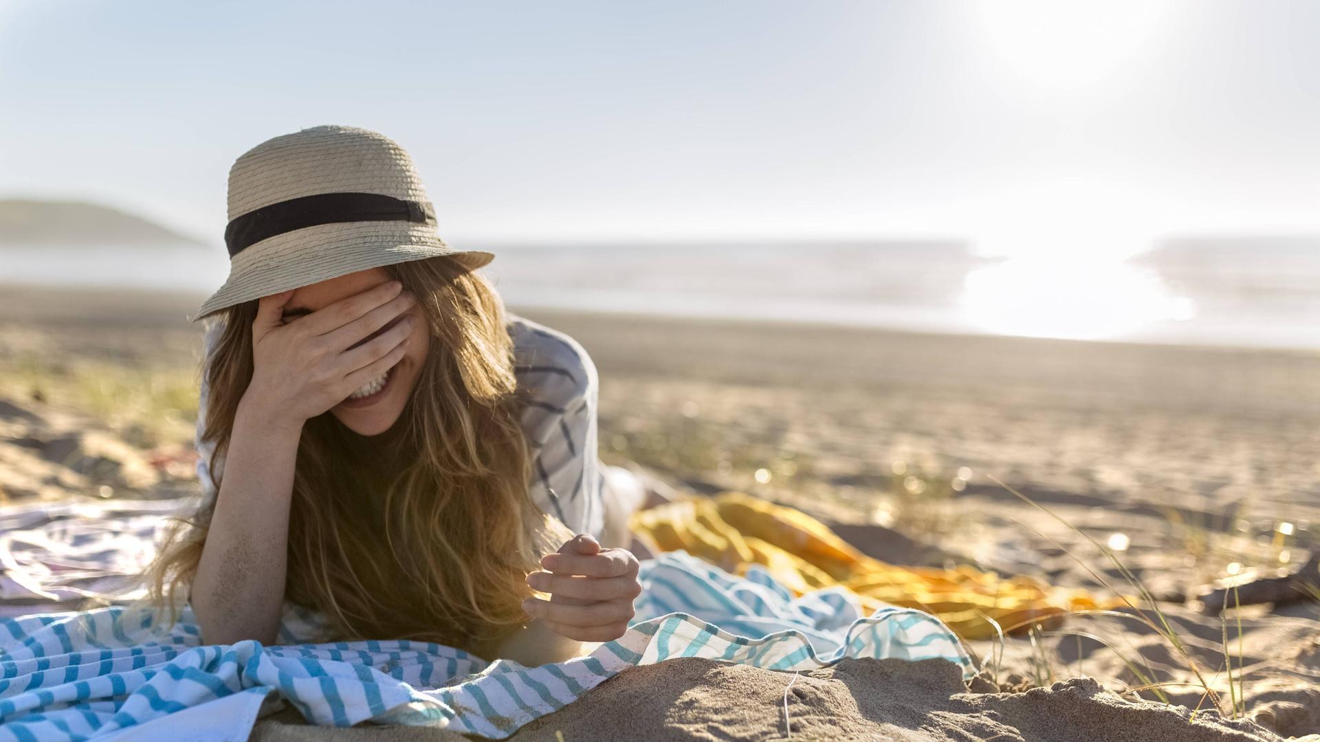 Eine Frau liegt mit dem Rücken zum Meer am Strand. Die Frau trägt einen Sonnenhut und eine Hemd und liegt auf ihrem Bauch auf einem Tuch. Sie hält ihre Hand vor ihre Augen und lacht.