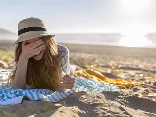 Eine Frau liegt mit dem Rücken zum Meer am Strand. Die Frau trägt einen Sonnenhut und eine Hemd und liegt auf ihrem Bauch auf einem Tuch. Sie hält ihre Hand vor ihre Augen und lacht. Eine Frau liegt mit dem Rücken zum Meer am Strand. Die Frau trägt einen Sonnenhut und eine Hemd und liegt auf ihrem Bauch auf einem Tuch. Sie hält ihre Hand vor ihre Augen und lacht.