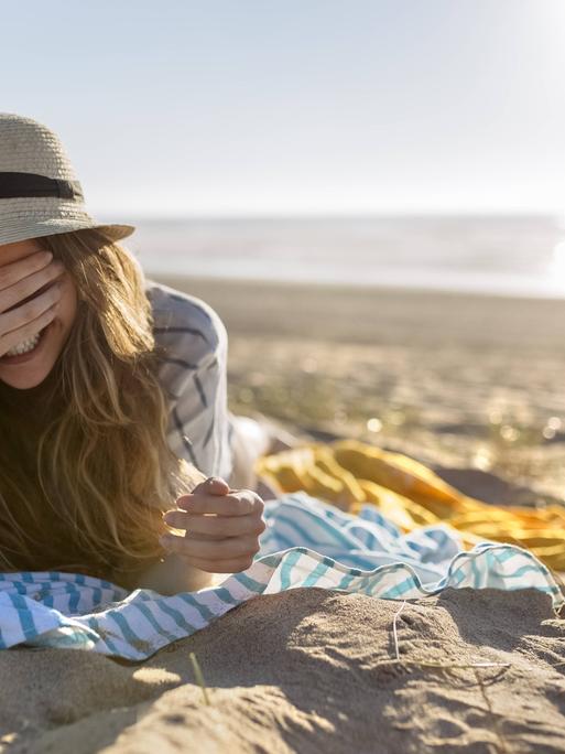 Eine Frau liegt mit dem Rücken zum Meer am Strand. Die Frau trägt einen Sonnenhut und eine Hemd und liegt auf ihrem Bauch auf einem Tuch. Sie hält ihre Hand vor ihre Augen und lacht. Eine Frau liegt mit dem Rücken zum Meer am Strand. Die Frau trägt einen Sonnenhut und eine Hemd und liegt auf ihrem Bauch auf einem Tuch. Sie hält ihre Hand vor ihre Augen und lacht.