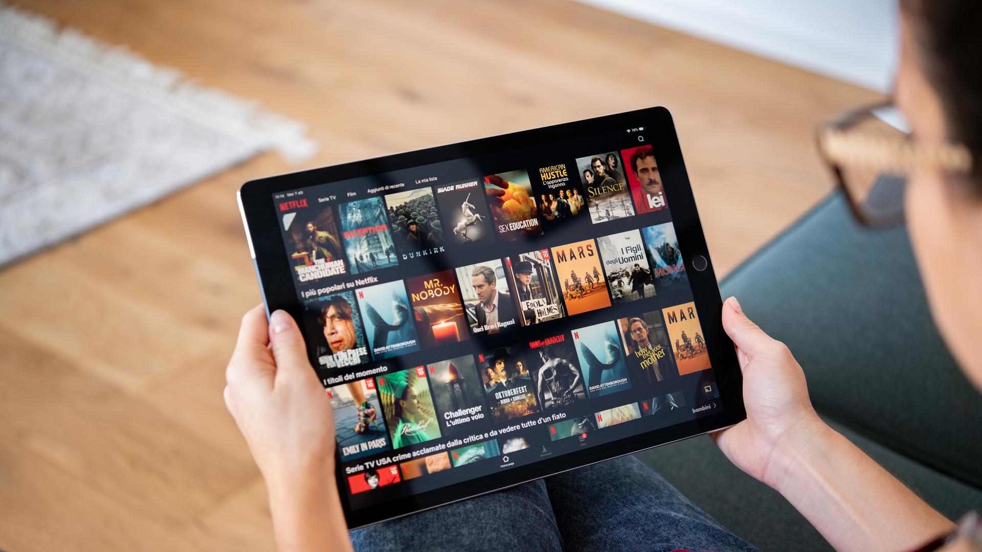 Eine Person sitzt auf dem Sofa und hält ein Tablet in der Hand. Sie ist damit beschäftigt, einen Film oder eine Serie auszuwählen. Eine Person sitzt auf dem Sofa und hält ein Tablet in der Hand. Sie ist damit beschäftigt, einen Film oder eine Serie auszuwählen.