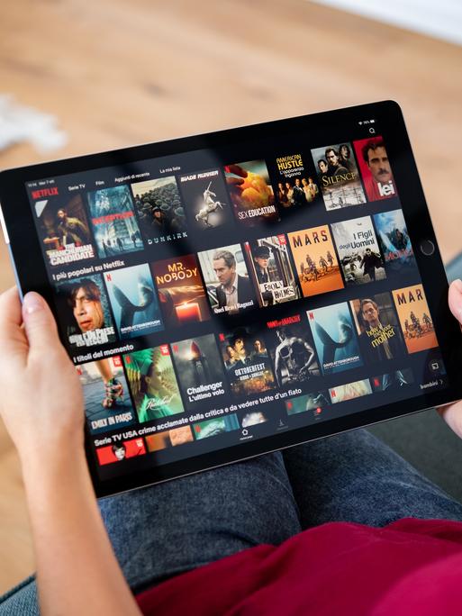 Eine Person sitzt auf dem Sofa und hält ein Tablet in der Hand. Sie ist damit beschäftigt, einen Film oder eine Serie auszuwählen.