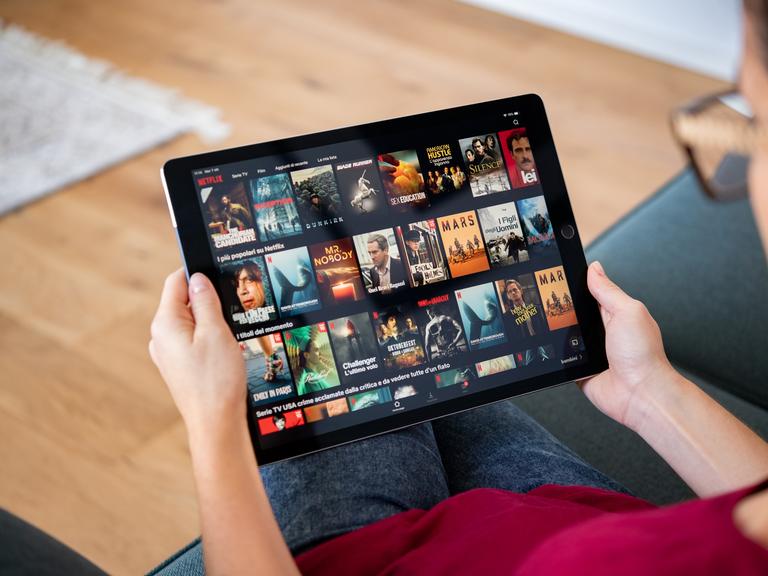 Eine Person sitzt auf dem Sofa und hält ein Tablet in der Hand. Sie ist damit beschäftigt, einen Film oder eine Serie auszuwählen.