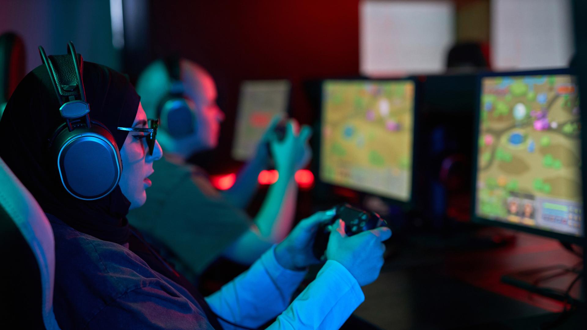 Seitenansicht-Porträt einer Frau, die einen Gaming-Controller hält und Videospiele im Neonlicht spielt. Neben ihr sitzt ein Mann, der auch spielt. Beide tragen Kopfhörer und sind konzentriert.