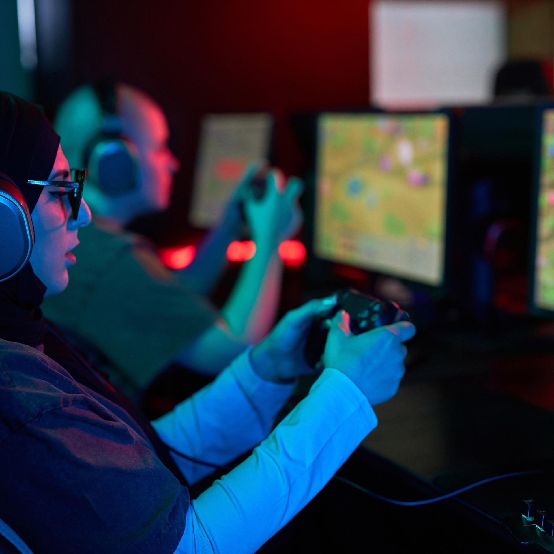 Seitenansicht-Porträt einer Frau, die einen Gaming-Controller hält und Videospiele im Neonlicht spielt. Neben ihr sitzt ein Mann, der auch spielt. Beide tragen Kopfhörer und sind konzentriert.
