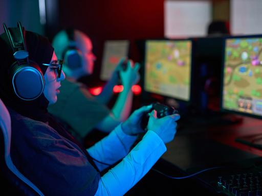 Seitenansicht-Porträt einer Frau, die einen Gaming-Controller hält und Videospiele im Neonlicht spielt. Neben ihr sitzt ein Mann, der auch spielt. Beide tragen Kopfhörer und sind konzentriert. Seitenansicht-Porträt einer Frau, die einen Gaming-Controller hält und Videospiele im Neonlicht spielt. Neben ihr sitzt ein Mann, der auch spielt. Beide tragen Kopfhörer und sind konzentriert.