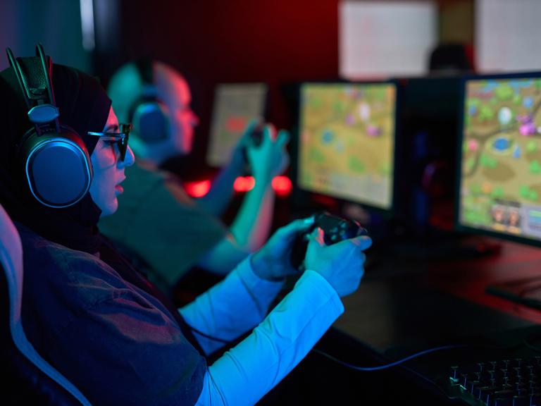 Seitenansicht-Porträt einer Frau, die einen Gaming-Controller hält und Videospiele im Neonlicht spielt. Neben ihr sitzt ein Mann, der auch spielt. Beide tragen Kopfhörer und sind konzentriert.