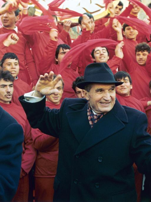 Der rumänische Diktator Nicolae Ceaușescu winkt am 24. November 1989 nach dem Parteitag in Bukarest der Bevölkerung zu. Im Hintergrund rot gekleidete junge Männer, die rote Tücher schwenken.