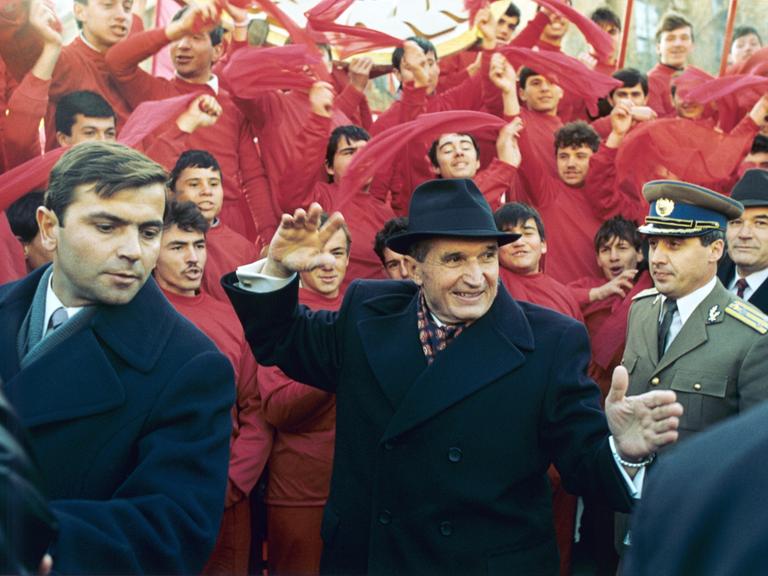 Der rumänische Diktator Nicolae Ceaușescu winkt am 24. November 1989 nach dem Parteitag in Bukarest der Bevölkerung zu. Im Hintergrund rot gekleidete junge Männer, die rote Tücher schwenken.