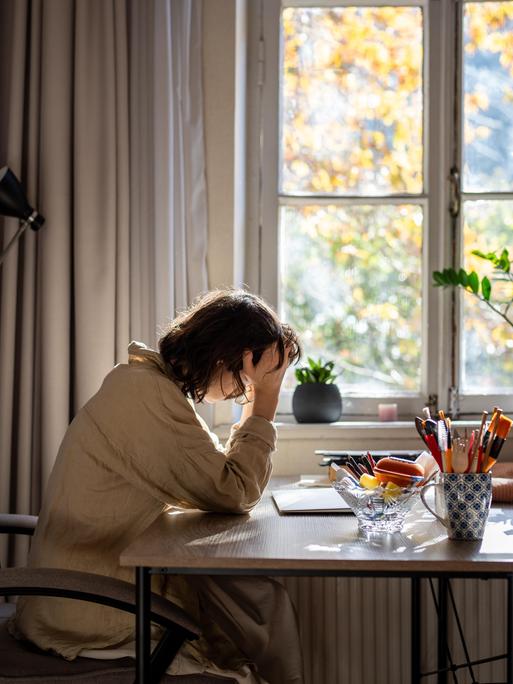 Eine offensichtlich verschlafene, halbwache Frau sitzt an einem Tisch und stützt den Kopf auf ihre rechte Hand