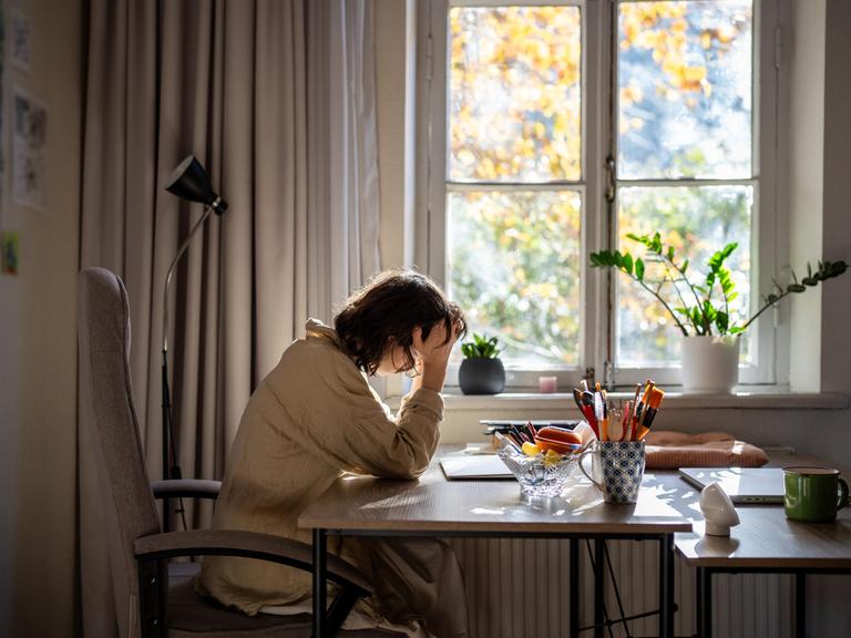 Eine offensichtlich verschlafene, halbwache Frau sitzt an einem Tisch und stützt den Kopf auf ihre rechte Hand