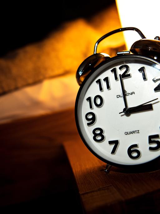 Ein Wecker steht neben einem Bett auf einem kleinen Tisch. Darauf steht auch eine Lampe, die warmes Licht macht. Der Wecker zeigt 3 Uhr.
