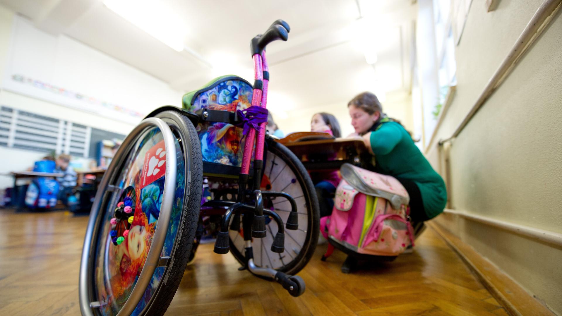 Ein Rollstuhl steht im Klassenzimmer, dahinter sitzen zwei Schülerinnen an einem Tisch Ein Rollstuhl steht im Klassenzimmer, dahinter sitzen zwei Schülerinnen an einem Tisch