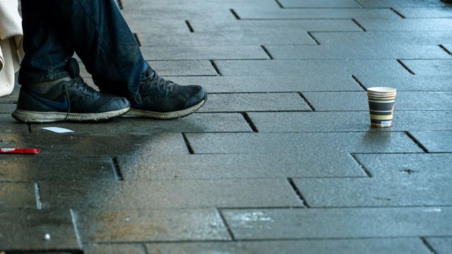 Schuhe eines Menschen, vor ihm steht ein Pappbecher