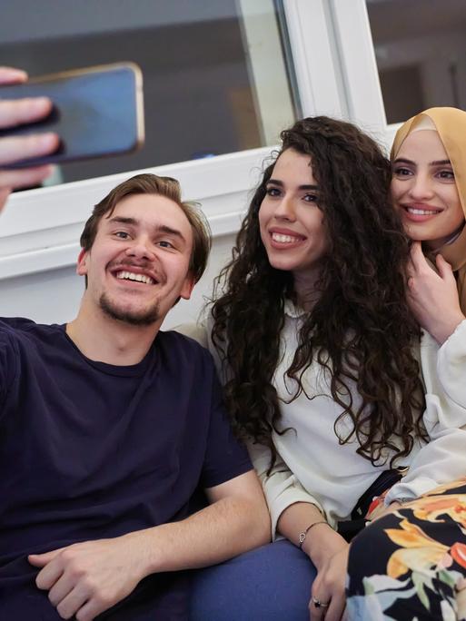 Drei Personen sitzen auf dem Sofa und machen ein Selfie. Die Frau (rechts im Bild) trägt ein Kopftuch als Zeichen ihrer Religiosität.