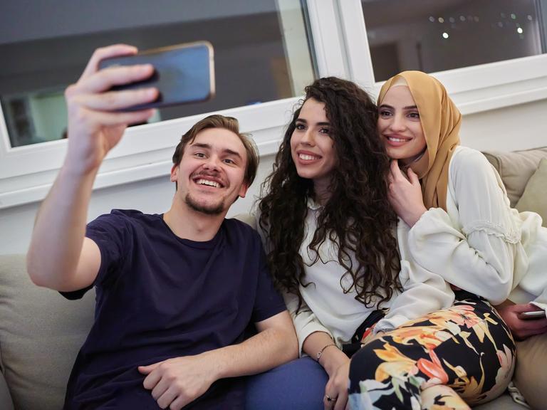 Drei Personen sitzen auf dem Sofa und machen ein Selfie. Die Frau (rechts im Bild) trägt ein Kopftuch als Zeichen ihrer Religiosität.