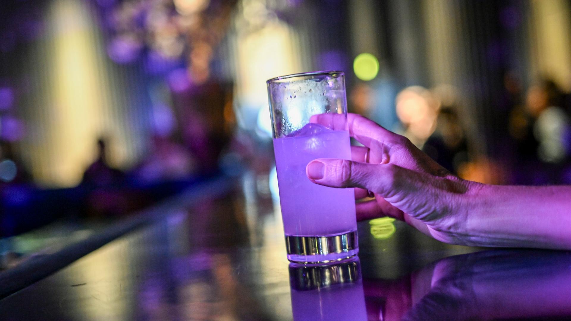 Eine Frau steht an einer Theke und greift nach einem Glas mit einem Cocktail, der violett leuchtet.