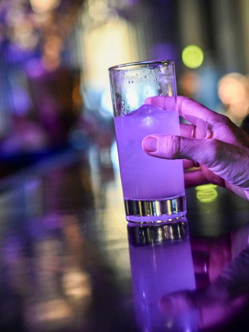 Eine Frau steht an einer Theke und greift nach einem Glas mit einem Cocktail, der violett leuchtet.