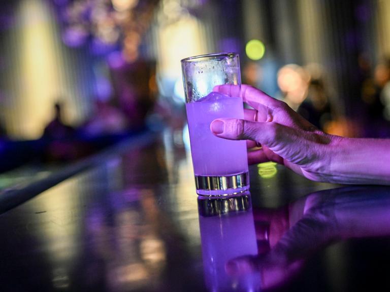 Eine Frau steht an einer Theke und greift nach einem Glas mit einem Cocktail, der violett leuchtet.