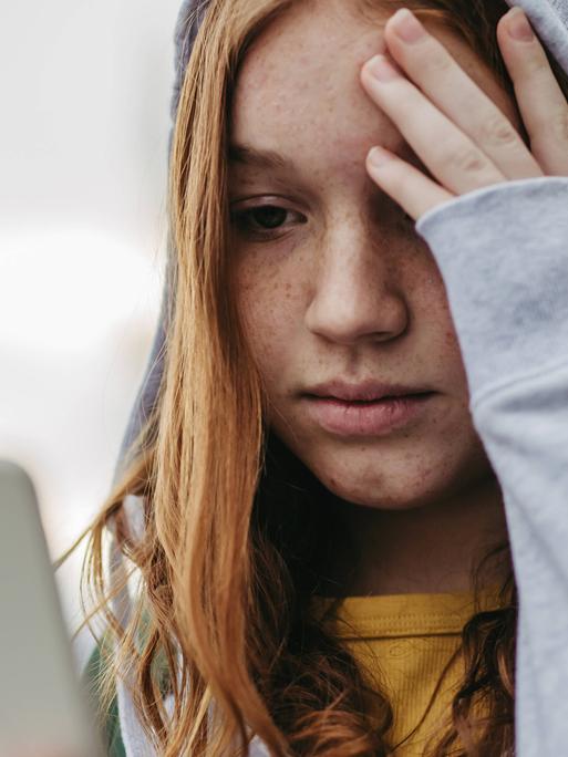 Foto einer jungen Frau, die traurig auf ihr Smartphone guckt und die Hand an die Stirn legt.