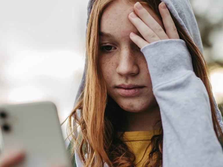 Foto einer jungen Frau, die traurig auf ihr Smartphone guckt und die Hand an die Stirn legt.