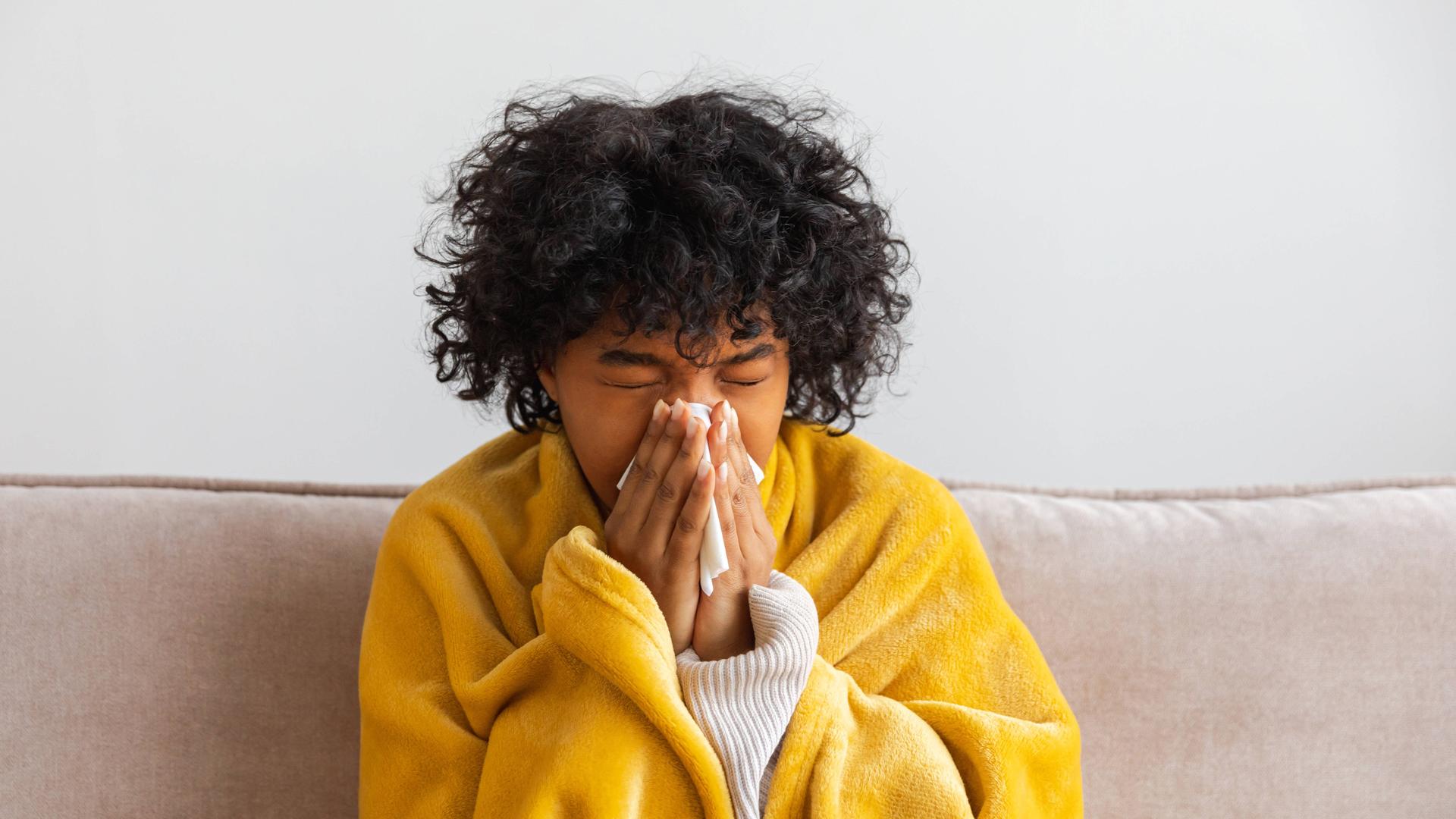 Unglückliche, erkrankte junge Afroamerikanerin mit Erkältung sitzt zu Hause auf dem Sofa. Mädchen bedeckt sich mit einer Decke und putzt sich die Nase mit einem Taschentuch.