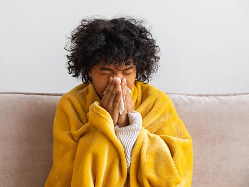 Unglückliche, erkrankte junge Afroamerikanerin mit Erkältung sitzt zu Hause auf dem Sofa. Mädchen bedeckt sich mit einer Decke und putzt sich die Nase mit einem Taschentuch. Unglückliche, erkrankte junge Afroamerikanerin mit Erkältung sitzt zu Hause auf dem Sofa. Mädchen bedeckt sich mit einer Decke und putzt sich die Nase mit einem Taschentuch.