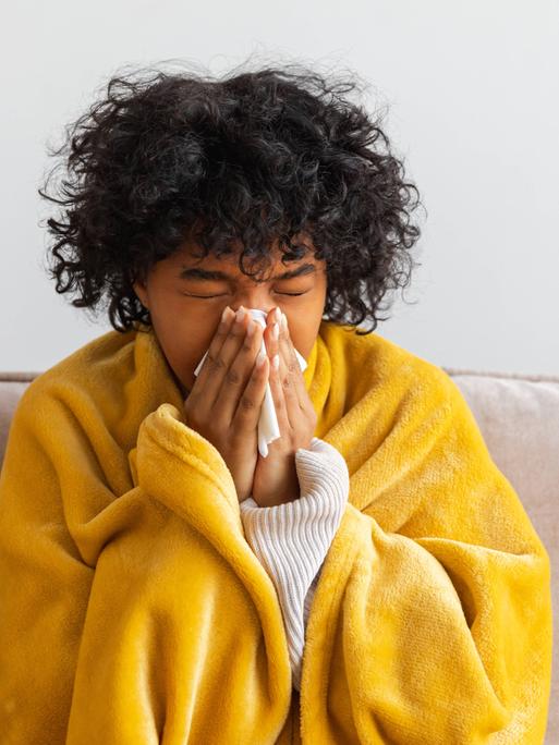 Unglückliche, erkrankte junge Afroamerikanerin mit Erkältung sitzt zu Hause auf dem Sofa. Mädchen bedeckt sich mit einer Decke und putzt sich die Nase mit einem Taschentuch. Unglückliche, erkrankte junge Afroamerikanerin mit Erkältung sitzt zu Hause auf dem Sofa. Mädchen bedeckt sich mit einer Decke und putzt sich die Nase mit einem Taschentuch.
