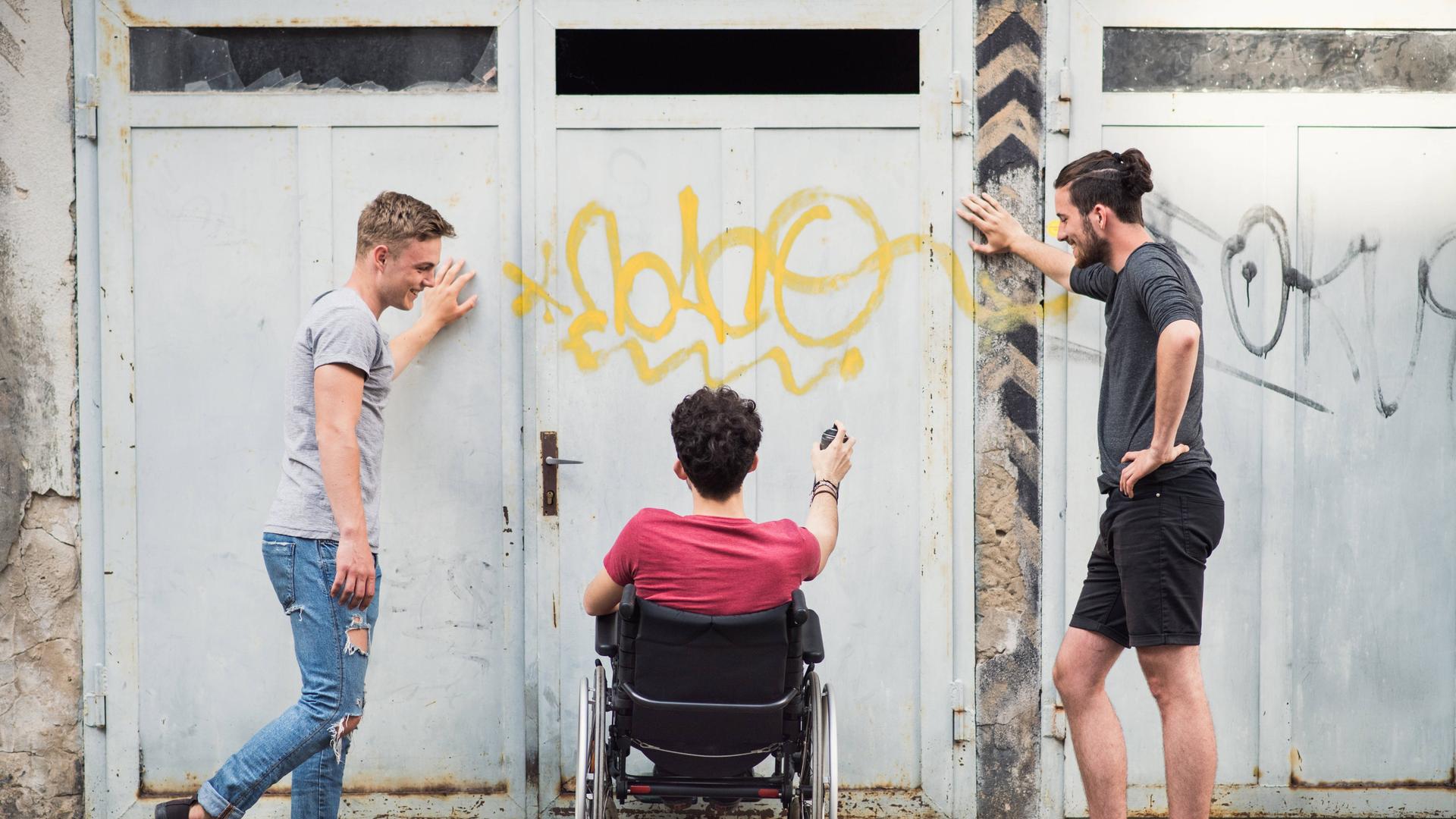 Drei Freunde beim Graffiti-Sprühen - zwei Männer stehen, einer sitzt im Rollstuhl Drei Freunde beim Graffiti-Sprühen - zwei Männer stehen, einer sitzt im Rollstuhl