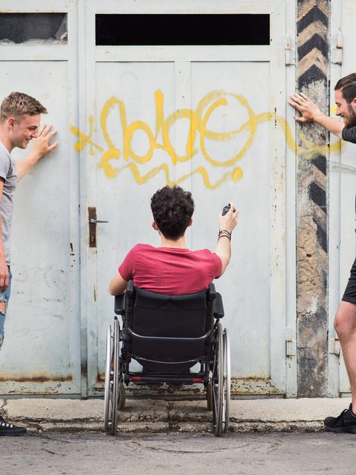 Drei Freunde beim Graffiti-Sprühen - zwei Männer stehen, einer sitzt im Rollstuhl