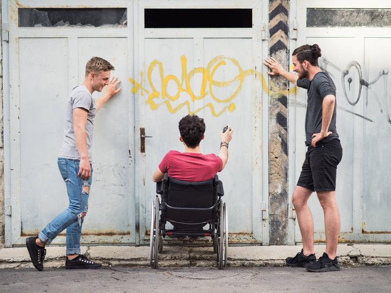 Drei Freunde beim Graffiti-Sprühen - zwei Männer stehen, einer sitzt im Rollstuhl
