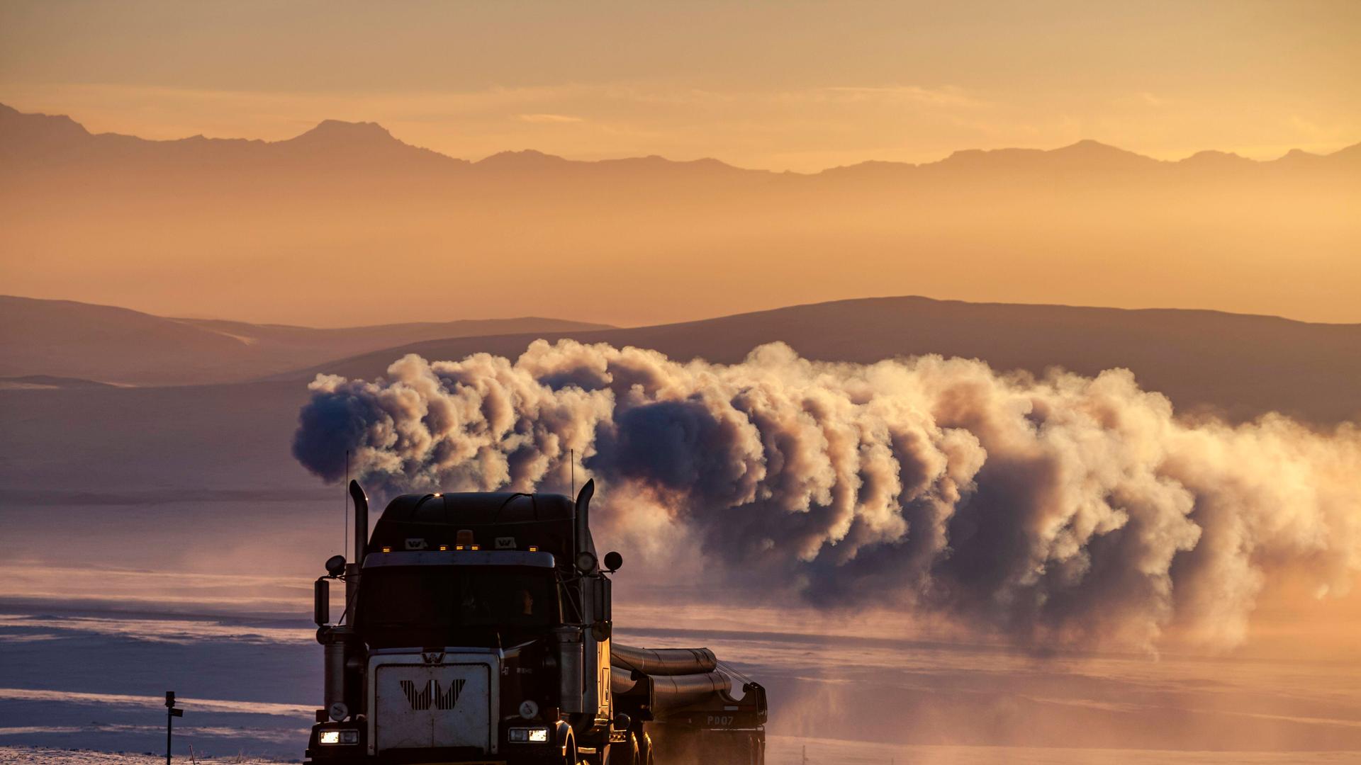 LKW auf verschneiter Piste mit großer Abgaswolke vor Bergen im Winter, Abendlicht, Dalton Highway, Alaska