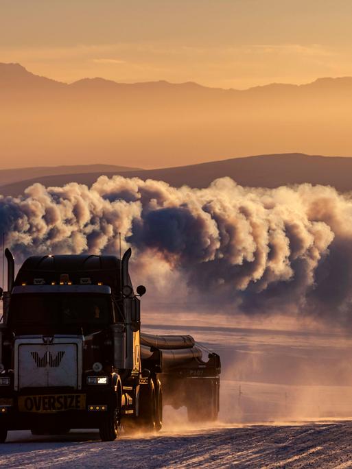LKW auf verschneiter Piste mit großer Abgaswolke vor Bergen im Winter, Abendlicht, Dalton Highway, Alaska