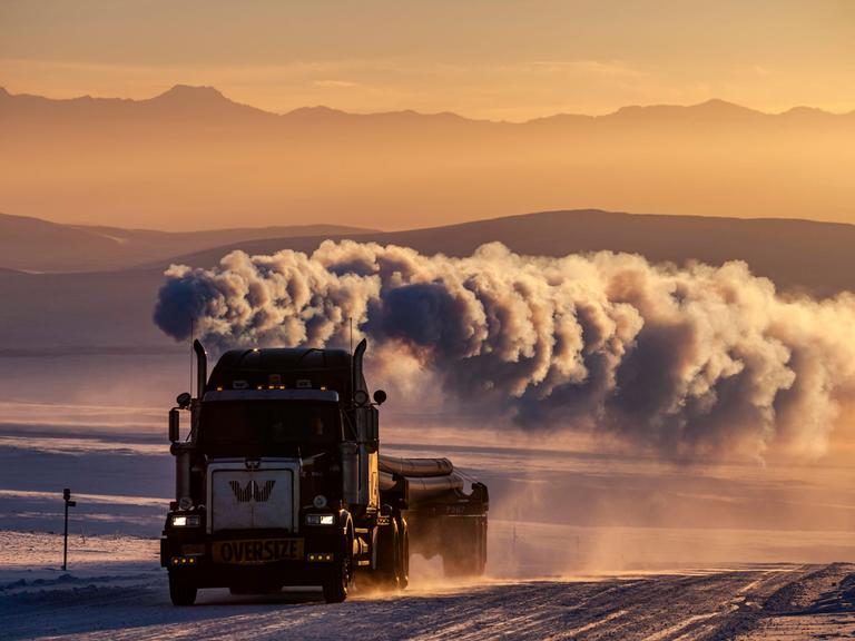 LKW auf verschneiter Piste mit großer Abgaswolke vor Bergen im Winter, Abendlicht, Dalton Highway, Alaska