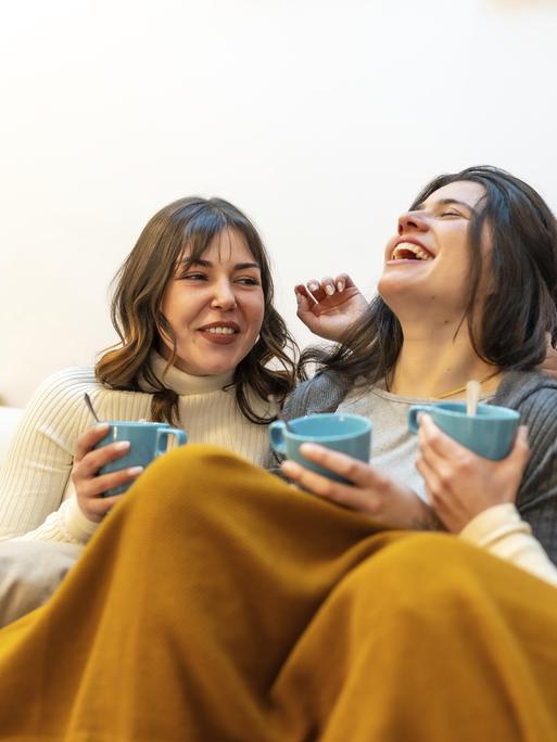 Glückliche Freundinnen, die zu Hause einen gemütlichen Moment zu dritt genießen, lachen und heiße Getränke trinken, während sie auf der Couch unter einer warmen Decke sitzen Glückliche Freundinnen, die zu Hause einen gemütlichen Moment zu dritt genießen, lachen und heiße Getränke trinken, während sie auf der Couch unter einer warmen Decke sitzen