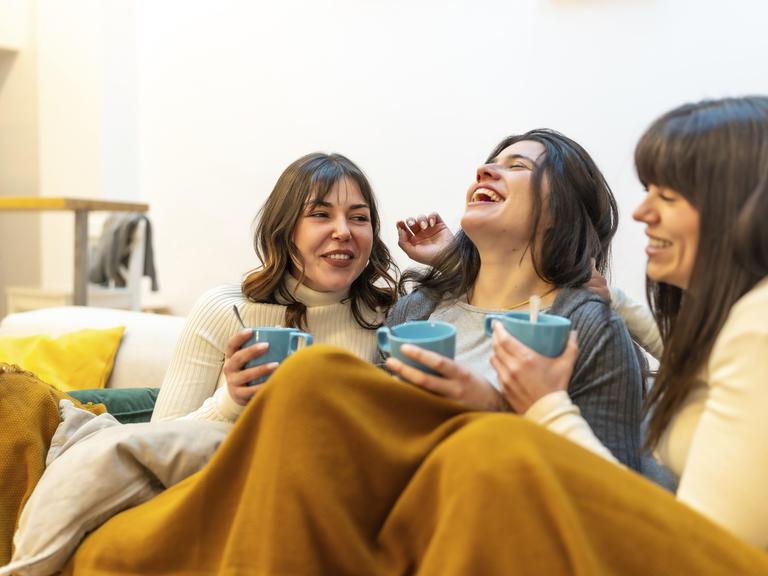 Glückliche Freundinnen, die zu Hause einen gemütlichen Moment zu dritt genießen, lachen und heiße Getränke trinken, während sie auf der Couch unter einer warmen Decke sitzen Glückliche Freundinnen, die zu Hause einen gemütlichen Moment zu dritt genießen, lachen und heiße Getränke trinken, während sie auf der Couch unter einer warmen Decke sitzen