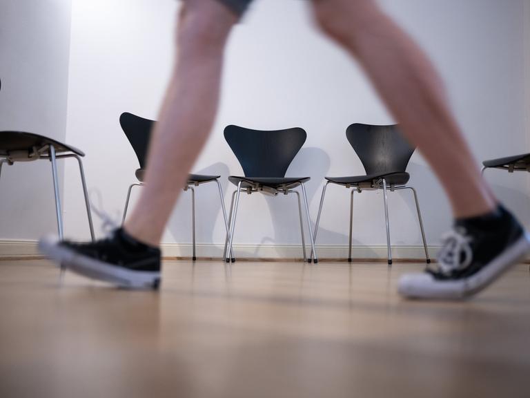 Sneaker tragende Person geht auf einem Gang an einer Reihe von Stühlen vorbei