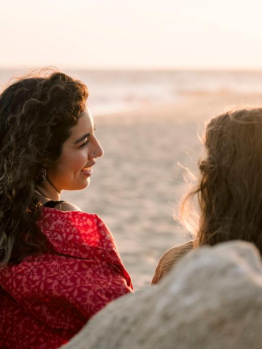 Zwei Frauen sitzen am Strand und unterhalten sich. Wir sehen sie von hinten.