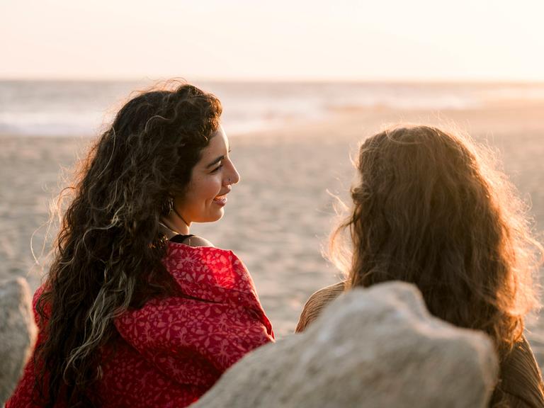 Zwei Frauen sitzen am Strand und unterhalten sich. Wir sehen sie von hinten.