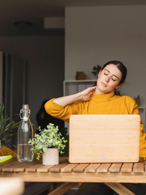 Eine Frau sitzt am Schreibtisch und berührt mit ihrer Hand ihren Nacken
