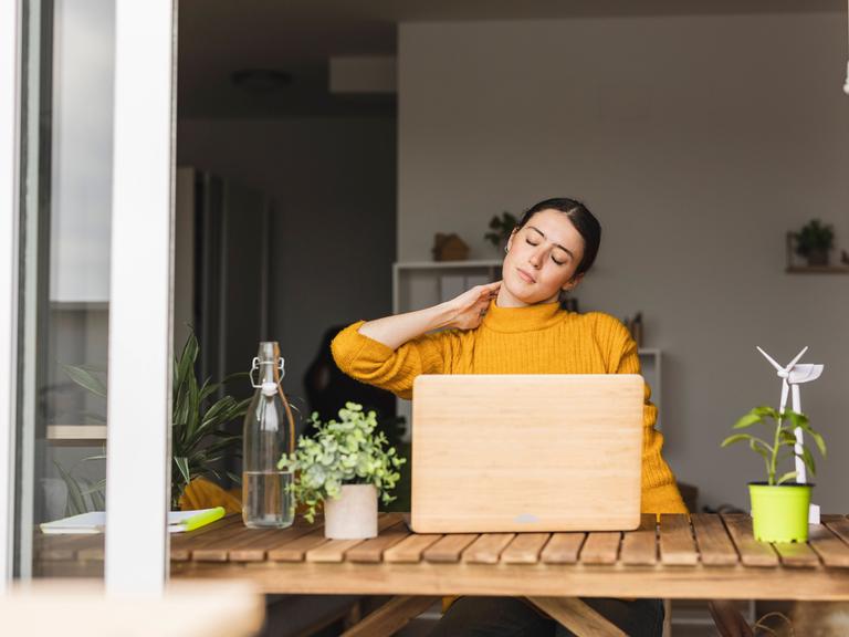 Eine Frau sitzt am Schreibtisch und berührt mit ihrer Hand ihren Nacken