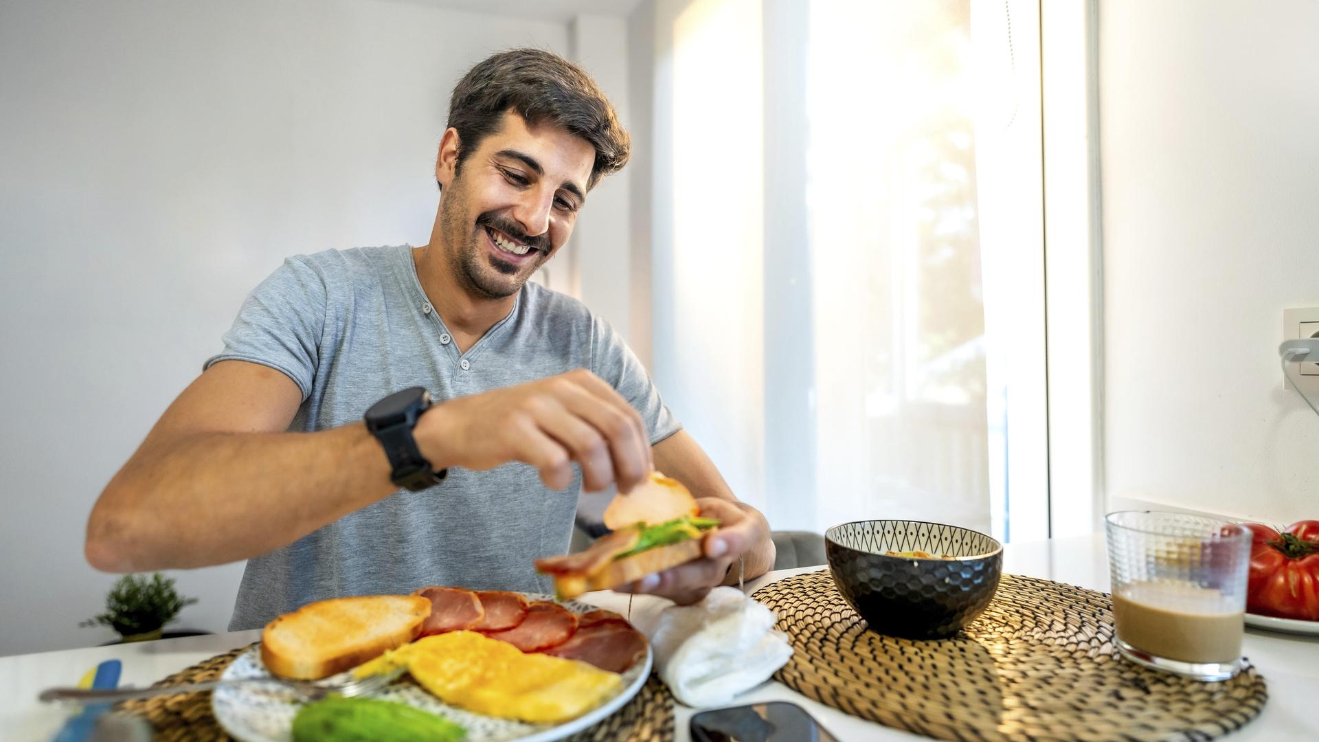 Ein Mann sitzt in der Küche am Frühstückstisch. Vor ihm stehen verschiedene Lebensmittel wie Obst, Brot, Müsli. Zu trinken hat er einen Milchkaffee. Der Mann belegt sich gerade ein Brot. Sein Handy liegt neben dem Teller.