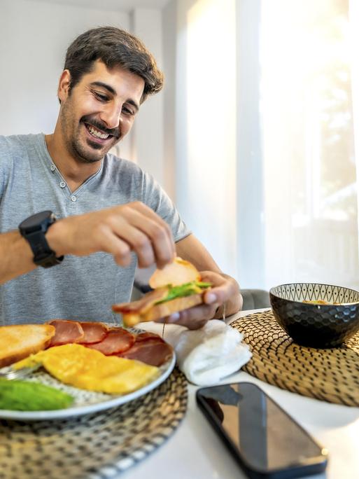 Ein Mann sitzt in der Küche am Frühstückstisch. Vor ihm stehen verschiedene Lebensmittel wie Obst, Brot, Müsli. Zu trinken hat er einen Milchkaffee. Der Mann belegt sich gerade ein Brot. Sein Handy liegt neben dem Teller. Ein Mann sitzt in der Küche am Frühstückstisch. Vor ihm stehen verschiedene Lebensmittel wie Obst, Brot, Müsli. Zu trinken hat er einen Milchkaffee. Der Mann belegt sich gerade ein Brot. Sein Handy liegt neben dem Teller.